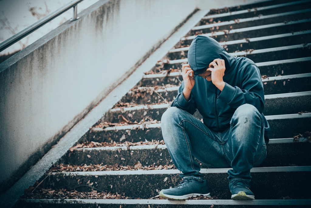 Man who is showing signs of meth use sits on staircase while hiding inside his own hoodie